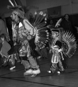 Man and young boy in full headdress leading a dance in a school. Photo by Larry Gouine.